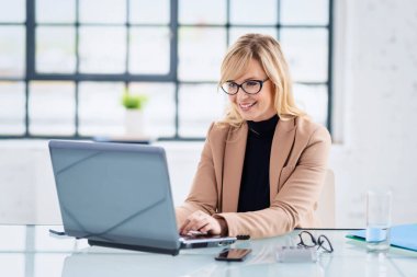 Dizüstü bilgisayarının arkasında oturmuş gülümseyen sarışın iş kadını fotoğrafı. Merkez Ofis.