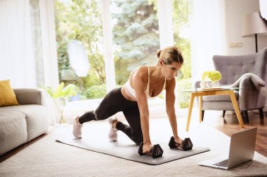 Evde vücut egzersizi yaparken bir kadının dambıl kullanması. Yoga minderinde egzersiz yaparken spor giyen ve dizüstü bilgisayar kullanan bir kadın. Sağlıklı yaşam.. 