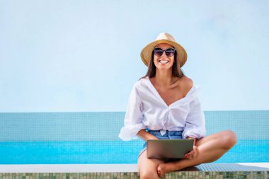 Hasır şapka ve güneş gözlüğü takan ve havuz başında dinlenirken dizüstü bilgisayar kullanan çekici bir kadının fotoğrafı.. 
