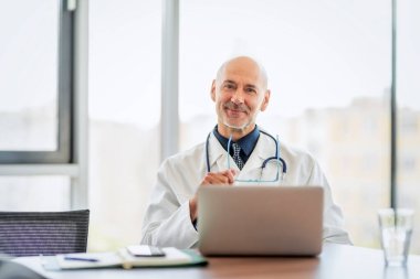 Male doctor looking at camera and smiling while sitting behind his laptop at doctors office.  