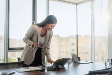 Happy businesswoman standing at office desk and using mobile phone and laptop while having video call. 