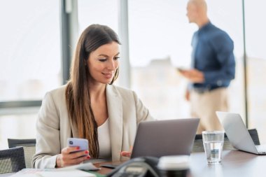Shot of confident businesswoman using her mobile phone and laptop while sitting at office desk and working. Unrecognizable businessman standing at the background. 