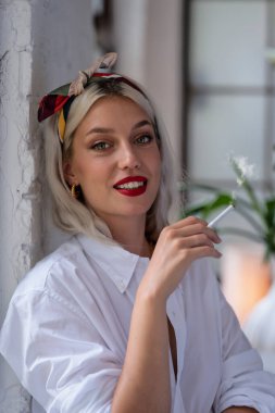 Portrait of beautiful green eyed young and blond haired woman wearing hair scarf and white shirt while smoking cigarette.