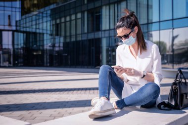 Mid adult woman using smartphone and walking outdoors in city during pandemia. Attractive female wearing face mask and plastic gloves. 