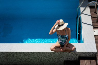 Happy woman wearing swimsuit and straw hat while relaxing at the poolside. 