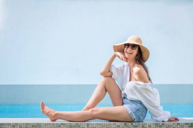 Happy woman relaxing by a swimming pool. Attractive female wearing straw hat and sunglasses while looking at camera and smiling. 