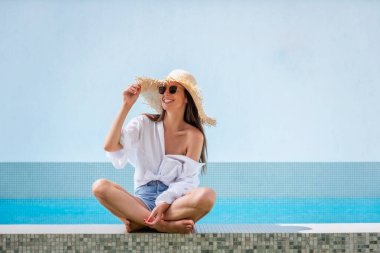 Happy woman relaxing by a swimming pool. Attractive female wearing straw hat and sunglasses while looking away and smiling. 
