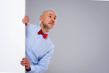 Studio shot of surprised face man looking up while standing behind an empty white board. Isolated grey background. Copy space. 