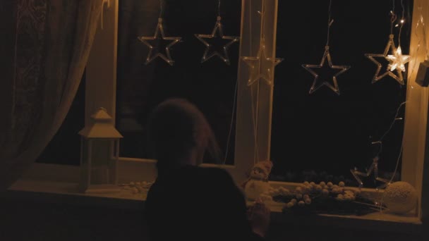 Девушка подходит к окну, где светит гирлянда — стоковое видео