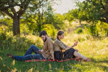 Bir çift yaz parkında çimenlikte oturmuş telefonlara bakıyor.