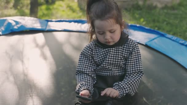 Lány nézi a telefont, miközben ül a trambulinon — Stock videók