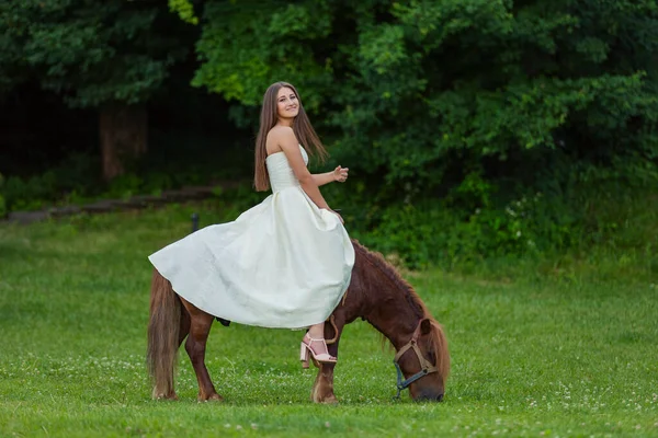 白いドレスの女の子が緑の芝生の上でポニーに乗って — ストック写真