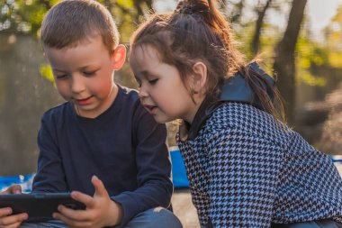 Çocuklar trambolinde oturup telefona bakıyorlar.