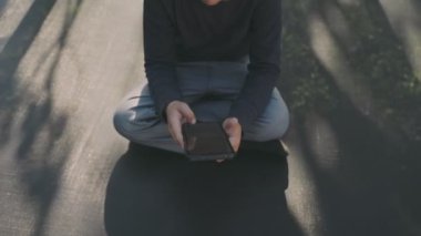 Küçük çocuk trambolinde otururken telefonla oynuyor.