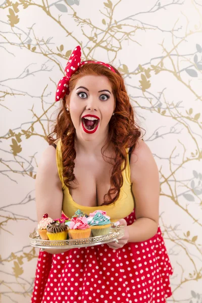 Beautiful pinup redhead girl holding a tray of colorful cupcakes — Stock Photo, Image