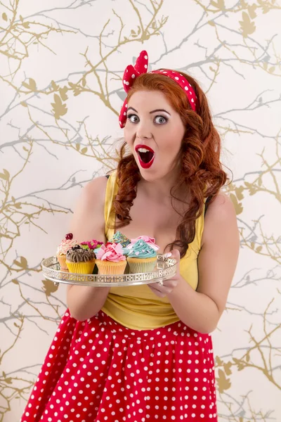 Menina ruiva pinup bonita segurando uma bandeja de cupcakes coloridos — Fotografia de Stock