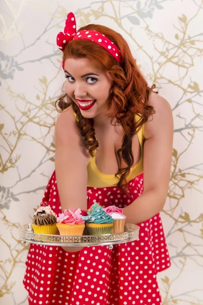 Hermosa chica pelirroja pinup sosteniendo una bandeja de cupcakes de colores — Foto de Stock