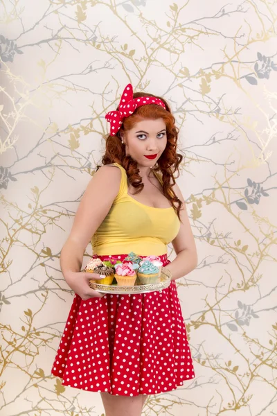 Beautiful pinup redhead girl holding a tray of colorful cupcakes — Stock Photo, Image