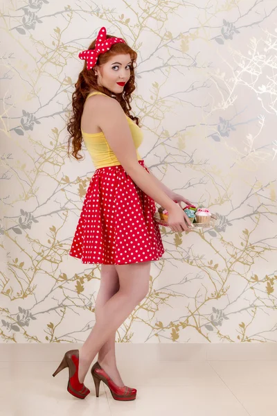 Hermosa chica pelirroja pinup sosteniendo una bandeja de cupcakes de colores — Foto de Stock