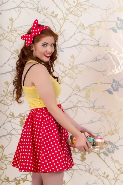 Beautiful pinup redhead girl holding a tray of colorful cupcakes — Stock Photo, Image