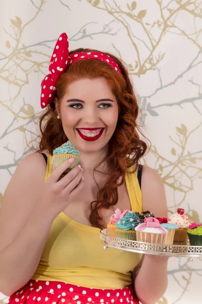 Hermosa chica pelirroja pinup sosteniendo una bandeja de cupcakes de colores — Foto de Stock