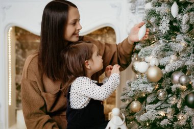 Güzel bir kızı olan genç bir anne şöminenin arka planında bir Noel ağacı süsler. Esmer, şirin bebeğinin Noel ağacı oyuncağını giymesini izlemekten zevk alıyor. Kış yeni yıl tatili