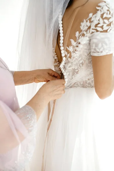 The bridesmaid helps the bride button up her white wedding dress on her wedding day. Mom\'s hands and buttons on the bride\'s dress close-up. An affectionate relationship between mother and daughter