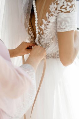 Nedime gelinin beyaz gelinliği iliklemesine yardım eder. Annenin elleri ve düğmeleri gelinin elbisesinde. Gelin gelinliğini giyer ve damatla tanışmaya hazırlanır.