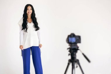 Bir kadın blogcu, beyaz bir fotoğraf stüdyosunda bir tripodun üzerinde profesyonel bir kamera ile video kaydediyor. Güzel genç esmer kadın gülümsüyor ve kameraya bakıyor.