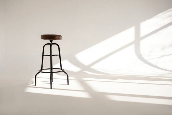 A white room with a shade from the sun through the window and one high bar stool. The style is minimalism in horizontal photography. Loneliness concept with one chair and white space