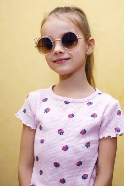 Baby girl in sunglasses on a yellow background. Portrait of a little girl in a vertical photo with round sunglasses. Cute baby posing in a t-shirt with a picture of berries