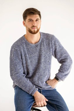 Portrait of a handsome man with a beard on a white background in a photo studio. A confident man in denim pants and a gray sweater sits on a chair. Vertical portrait of a 30 year old man