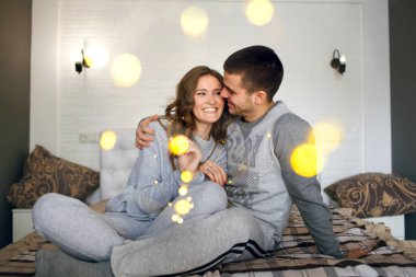 A couple in love in home clothes sits on the bed and hugs, throwing a garland of yellow lights over themselves. Happy man and young woman have fun decorating their home before christmas
