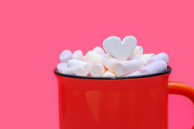 Çok renkli lokum ve kalp şeklinde şekerlemelerle süslenmiş büyük bir kupa sıcak kakao. Pembe arka planda kırmızı bardakta marşmelovlu leziz bir sıcak içecek. Sevgililer Günü konsepti