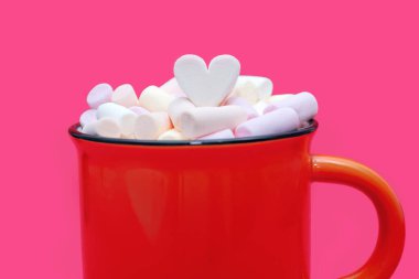 Kalp şeklindeki şekerlemelerle süslenmiş, çok renkli şekerlemeli büyük bir fincan sıcak kakao. Pembe arka planda turuncu bardakta marşmelovlu lezzetli bir sıcak içecek.