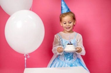 Pembe arka planda beyaz balonlu mavi elbiseli bir kız doğum gününde mumlu bir pasta tutuyor. Çocuk doğum gününü kutluyor ve mumu söndürmeye hazırlanırken gülümsüyor.