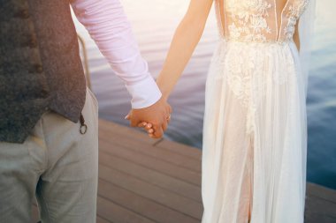 Gelinlik giymiş yeni evliler ırmağın arka planında el ele tutuşuyorlar. Gelin ve damat gün batımında birbirlerine sarılarak iskelede yürürler. Yeni evlilerin elleri. Yakın plan.