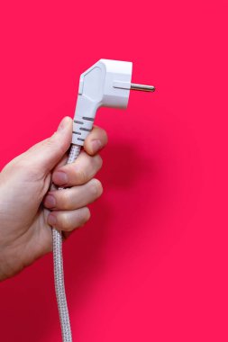 A man's hand holds an electric plug for an outlet on a pink background. The concept of electricity and its importance in everyday life. Electrical plug without socket