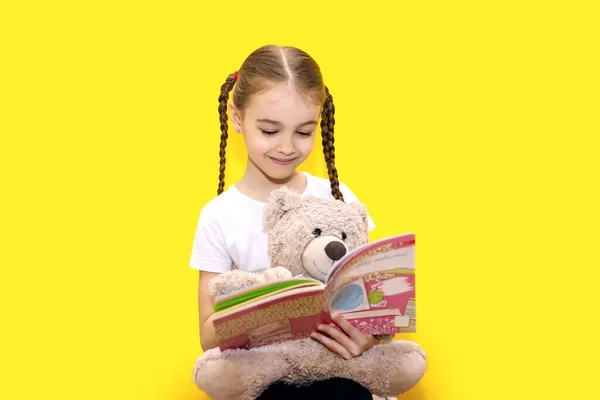 Girl Child Reads Book Holds Teddy Bear Yellow Background Cute — Photo