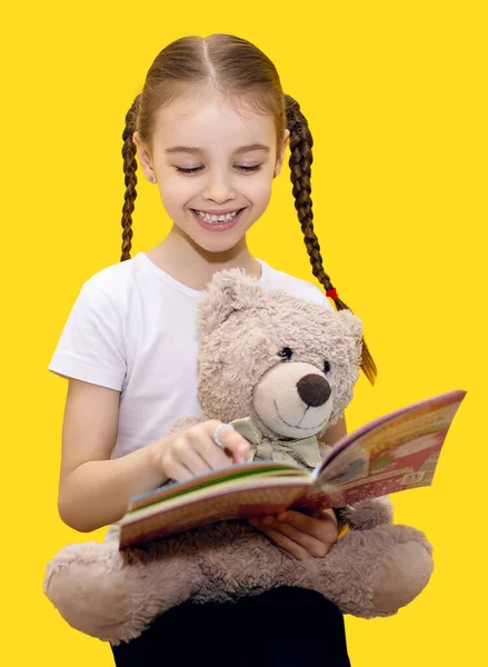 女の子の子供は本を読み 黄色の背景にテディベアを保持します 可愛い子がテディベアで読み書きや遊びを学んでいます 垂直写真の概念を学ぶ子供たち — ストック写真