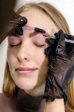 Kaş ustası bir takım aletler kullanıyor tarakları tarıyor ve müşterinin derisine cımbızla yaklaşan istenmeyen tüyleri alıyor. Kızıl saçlı, çilli, şekilli ve dikey bir fotoğrafta kaşlarını boyayan bir kız.