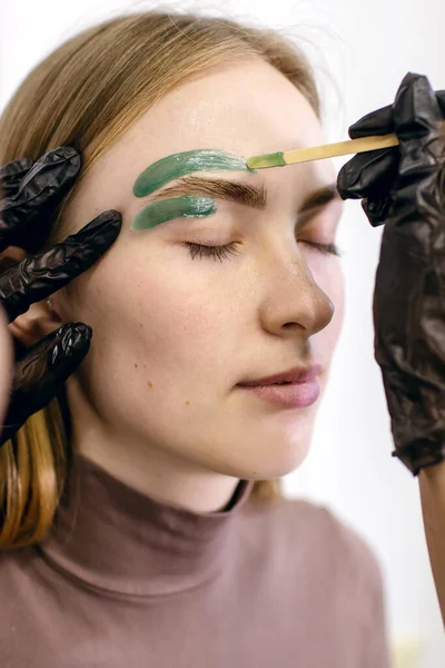 The process of removing hair around the eyebrows with a special resin or green wax on the skin of a red-haired girl with freckles. Eyebrow shaping and coloring in a beauty salon