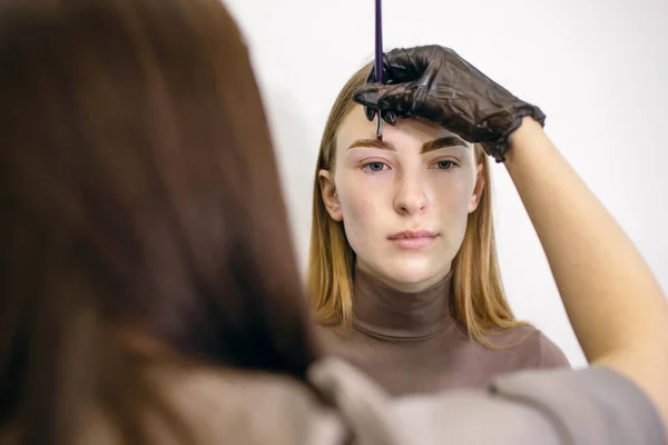 Eyebrow master shapes and paints with a brush the eyebrows of a red-haired girl with freckles. Young woman in the process of dyeing eyebrows and the work of a master stylist in a beauty salon