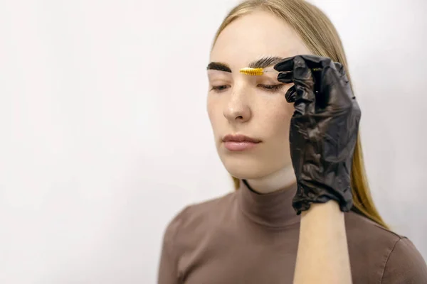 Eyebrow master shapes and paints with a brush the eyebrows of a red-haired girl with freckles in a beauty salon. Portrait of a young woman in the process of dyeing her eyebrows. Free space for text