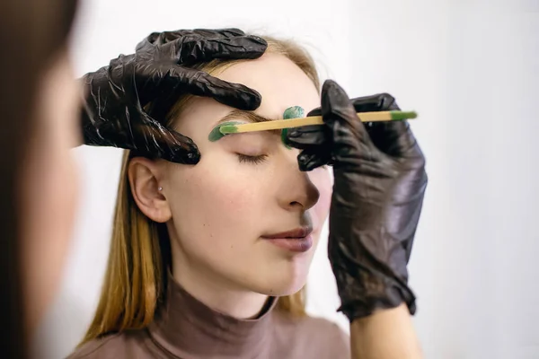 The process of removing hair around the eyebrows with a special resin or green wax on the skin of a red-haired girl with freckles. Eyebrow correction and coloring in a beauty salon