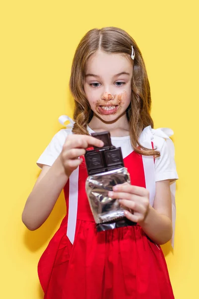 A child girl in a red dress holds a chocolate bar in her hands on a yellow background. Funny child stained his face with chocolate. The concept of children\'s love for sweets