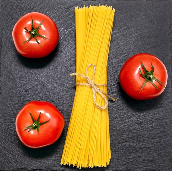 Siyah kaba bir arka planda çiğ spagetti ve kırmızı domatesler. Vejetaryen yemeği hazırlamak için ürünler. Yukarıdan güzel bir çiğ yemek modeli. Üç kırmızı domates ve uzun spagetti.