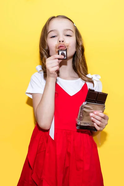 A little girl eats a chocolate bar and enjoys on a yellow background. A child in a red dress stained his face with chocolate. The concept of children\'s love for sweets