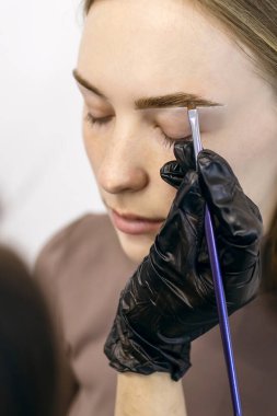 Kaş ustası, bir güzellik salonunda yakın plan kızıl saçlı bir kızın kaşlarını fırçaladı ve boyadı. Dikey bir arka plana kaşlarını boyama sürecinde olan genç bir kadın.
