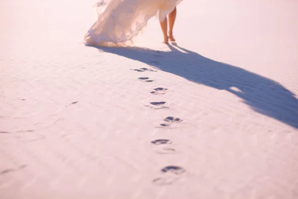A bride in a wedding dress walks in the desert on the sand. Footprints in the sand from the feet of a slender bride. Beautiful sunset and woman\'s feet with focus on footprints in the sand